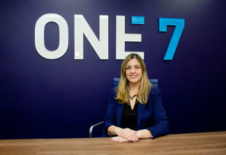 Patricia Baceti sentada com as maos em uma mesa e o logo da One7 ao fundo